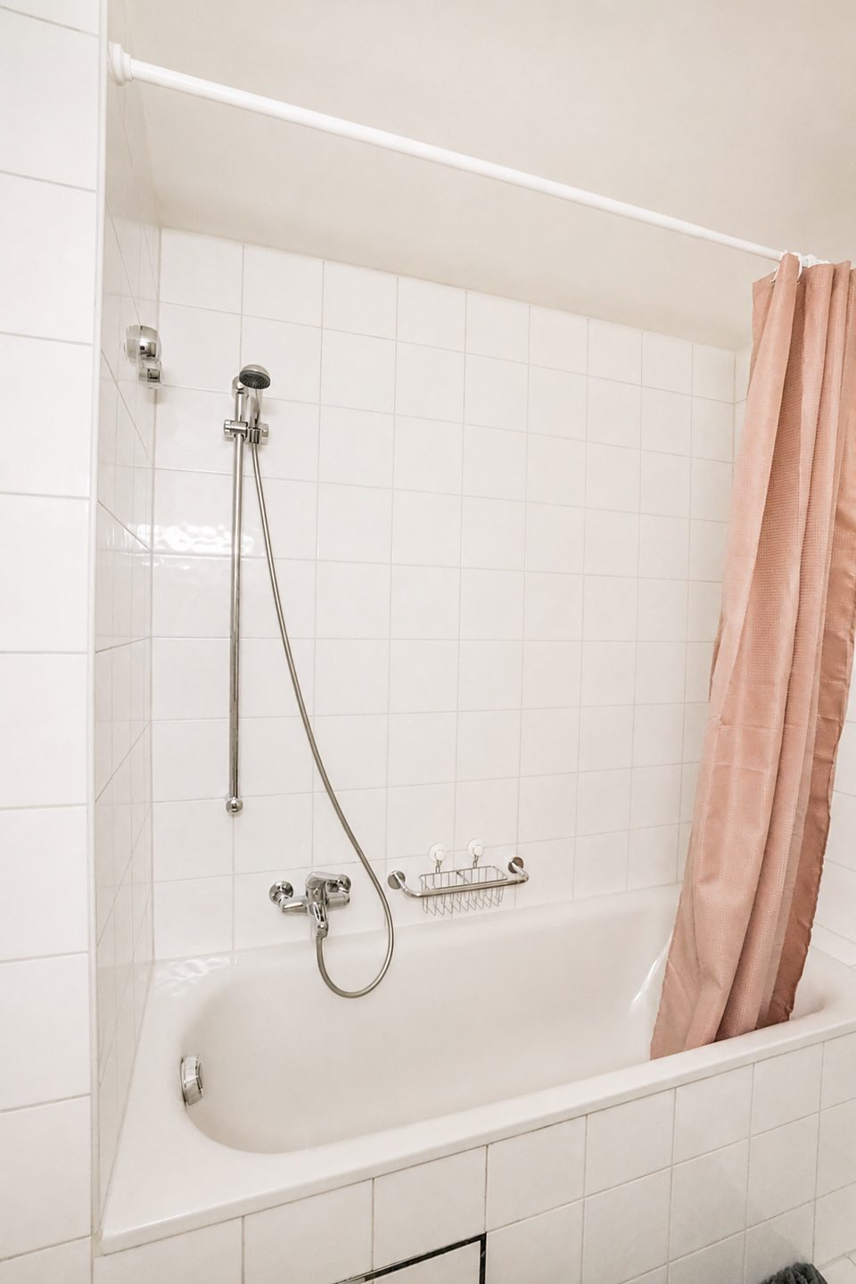 White-tiled bathtub with a handheld shower on a rail and a soft pink shower curtain.