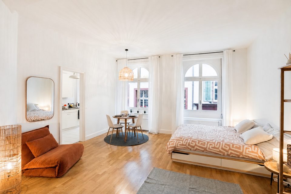 The studio in daylight: a double bed, a rust-coloured sofa and a round dining table beneath two tall arched windows.