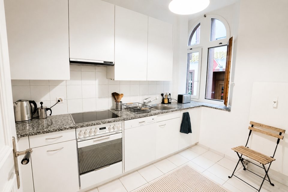 White kitchen with a ceramic hob, oven, sink and granite worktop beside an arched window.