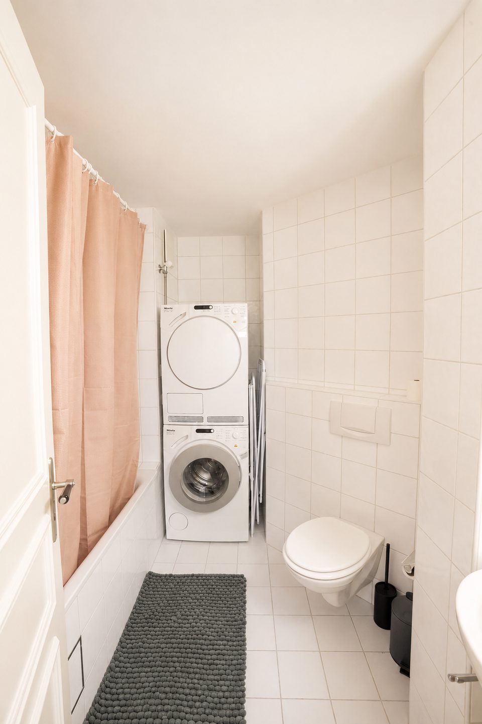 Stacked washing machine and dryer at the end of the tiled bathroom, beside the bathtub and toilet.