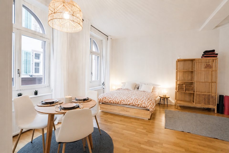 Round dining table set for three by an arched window, with the double bed and a slatted wooden wardrobe beyond.